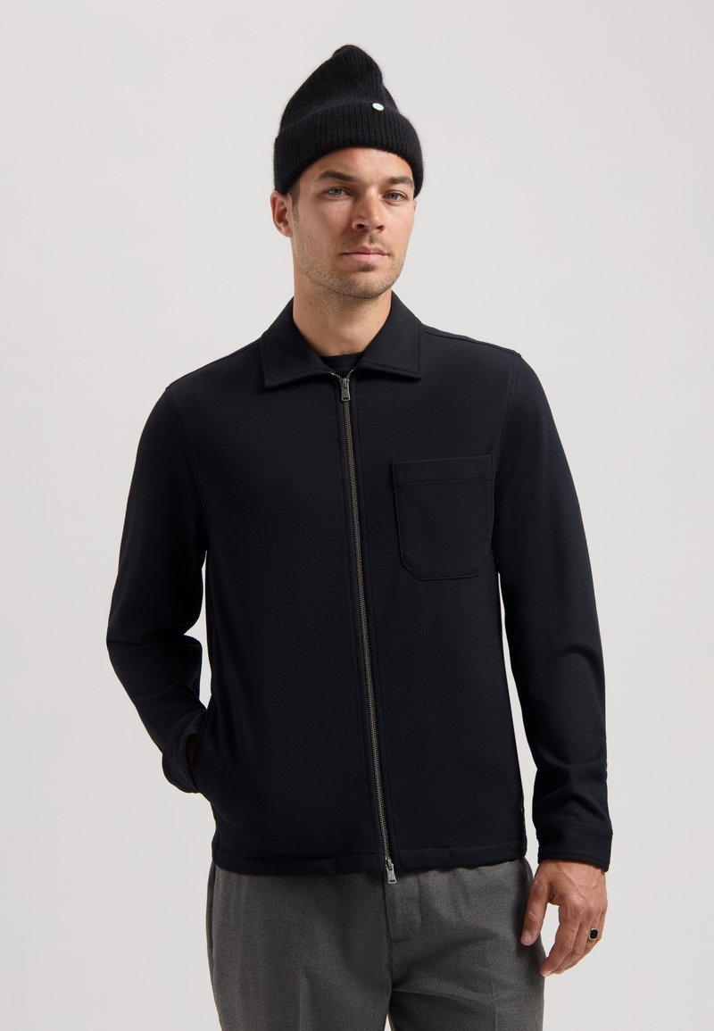 Chaqueta negra con cremallera, cuello, mangas largas y un bolsillo en el pecho. Textura suave, combinada con pantalones grises y un gorro de punto negro.