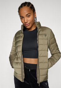 Veste matelassée vert olive avec un col haut, fermeture éclair et coutures horizontales. Portée par-dessus un top noir court, avec des accents de marque subtils.