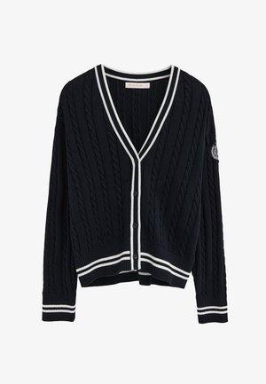 Cardigan en maille torsadée bleu marine avec bordure blanche sur le col, les poignets et l'ourlet, fermeture à boutons, et écusson brodé sur la manche gauche.