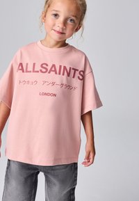 Vaaleanpunainen ylipitkä t-paita, jossa on tummanpunainen "ALLSAINTS"-logo, lyhyet hihat ja pyöreä pääntie, yhdistetty harmaisiin farkkuihin.