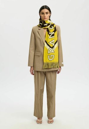 Vrouw draagt een beige pak en open teen hakken, geaccentueerd met een grote geel-zwarte sjaal met de tekst "GRANDE" om haar nek gewikkeld.