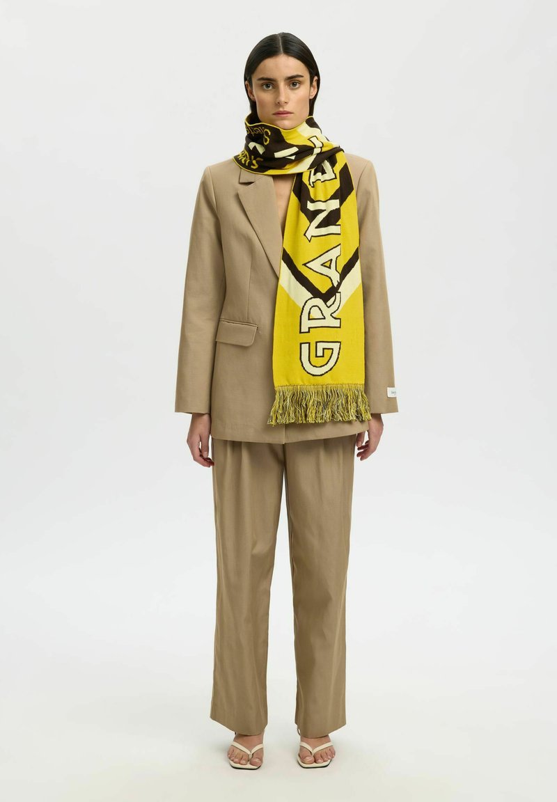 Femme portant un costume beige et des talons ouverts, accessoirisée d'une grande écharpe jaune et noire portant la mention "GRANDE" enroulée autour de son cou.