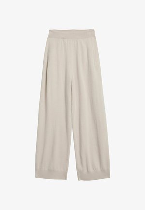 Kobieta stoi boso, ubrana w beżowy bezrękawnik i pasujące szerokie dziergane spodnie na jednolitym tle.