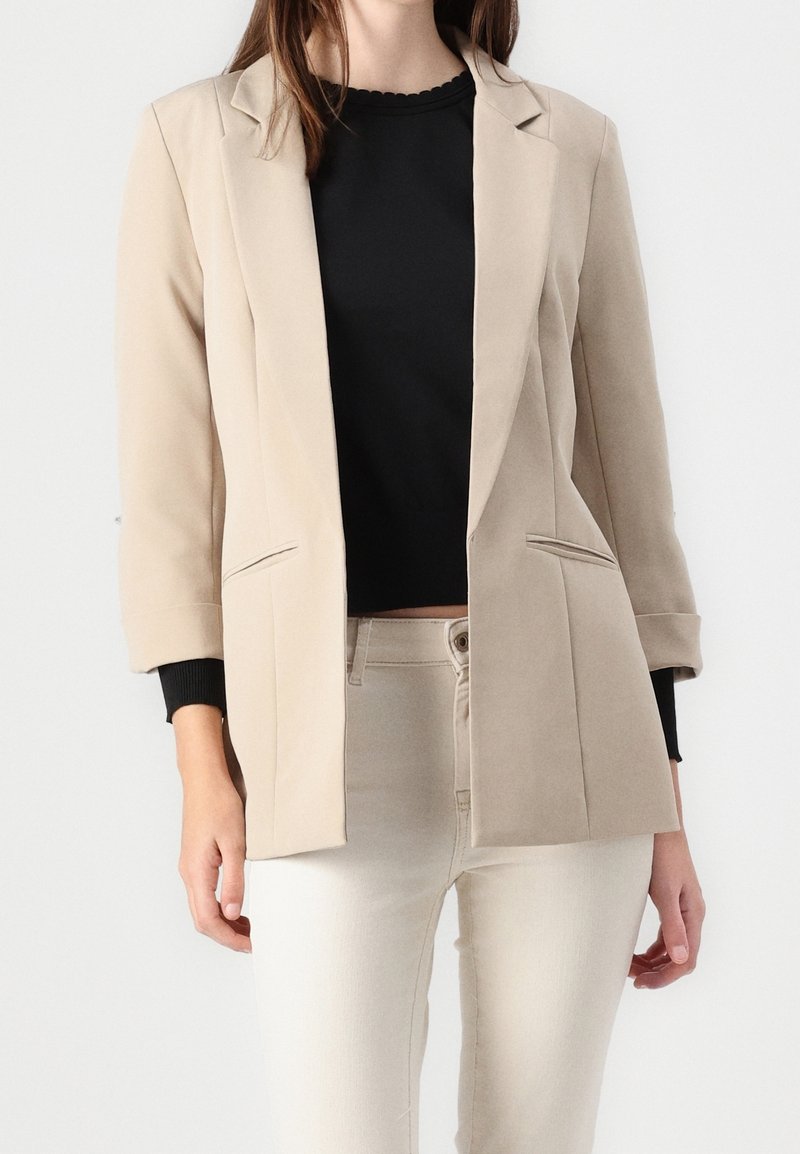 Femme portant un blazer beige sur un top noir et un pantalon clair, debout devant un fond blanc uni.