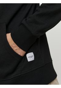 Černá mikina vyrobená z měkké tkaniny, s pružnými manžetami, přední kapsou a bílou nášivkou s nápisem "JACK & JONES EST. 1990" na boku.