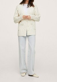 Woman wearing a cream cable-knit cardigan, white top, light blue wide-leg jeans, and cream ballet flats standing against a neutral background.