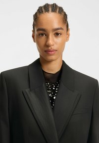 Zwarte blazer met satijnen revers, enkele borstzak, gecombineerd met een zwarte top met pailletten. Glad materiaal met een getailleerde pasvorm.