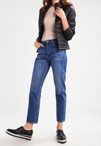 Sort dunjakke, beige ribbet sweater, blå high-waisted jeans og sorte platform sko med hvide striber, stående mod en lys baggrund.