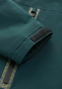Tissu de veste vert foncé avec un poignet à fermeture Velcro, des accents de couture jaune et une fermeture éclair noire sous un superposé géométrique.