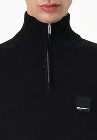Maglione nero a maglia con zip a un quarto, caratterizzato da un alto colletto a coste, una zip argentata e un'etichetta nera squadrata con il testo "KARL LAGERFELD JEANS".