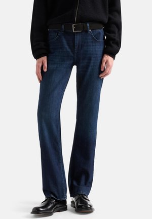 Person trägt dunkelblaue Jeans mit geradem Bein, schwarzen Ledergürtel, schwarze Jacke und schwarze polierte elegante Schuhe und steht vor weißem Hintergrund.