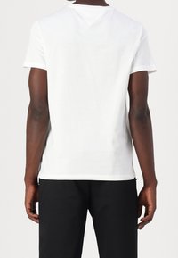 Biała bawełniana koszulka z krótkim rękawem, okrągłym dekoltem i delikatnym detalem w kształcie litery V na plecach, zestawiona z czarnymi spodniami.