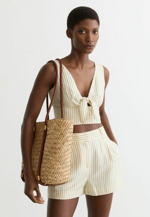 Femeie purtând un top crop crem cu dungi subțiri și fundă în față, și pantaloni scurți cu talie înaltă asortați, purtând o geantă mare împletită cu mânere împletite.