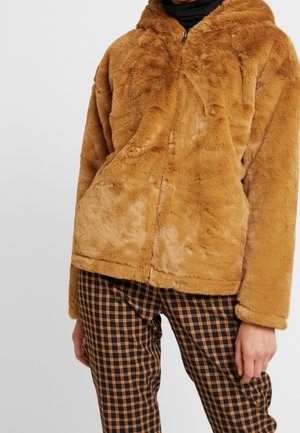 Personne portant une veste zippée en fausse fourrure couleur sable sur un pantalon à carreaux noir et orange, debout devant un fond clair uni.