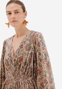Šaty s květinovým vzorem z jemného materiálu, s hlubokým výstřihem do V a smockovaným pasem. Velké náušnice ve tvaru pomerančové květiny doplňují celkový vzhled.