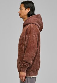 Felpa oversized marrone con finitura testurizzata, cappuccio con coulisse e tasca frontale. Il tessuto appare morbido e usurato.