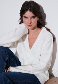 Cardigan alb pufos cu decolteu în V, mâneci lungi și închidere cu un singur buton, purtat cu blugi din denim de culoare bleumarin. Textură netedă și design casual.