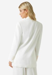 Hvid skræddersyet blazer med lange ærmer, glat stof, enkelt slids bagpå og manchetter med knapper, der fremhæver et rent og struktureret design.