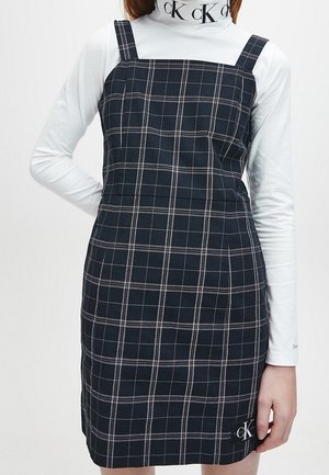 Vestido sin mangas a cuadros en blanco y negro, llevado sobre un cuello vuelto blanco de manga larga con el logo "CK" en el cuello y el dobladillo del vestido.