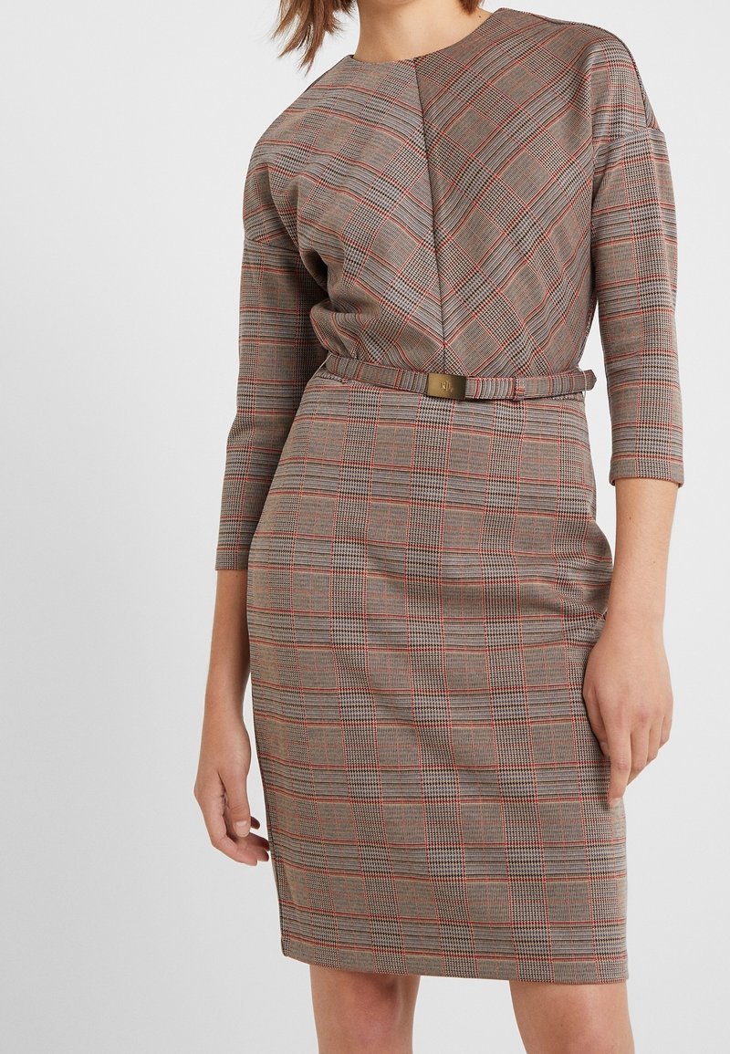 Femme portant une robe ajustée à carreaux longueur genou, manches trois-quarts et fine ceinture à la taille.