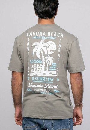 Homme portant un t-shirt gris avec un motif palmier et océan blanc et bleu de Laguna Beach ainsi qu'un texte au dos, associé à un jean bleu.