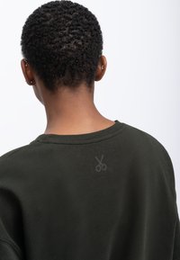 Donkergroene sweatshirt met een ribbelboord aan de halslijn, voorzien van een subtiele schaarafdruk op de achterkant. Gemaakt van gestructureerde stof.