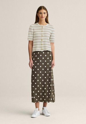 Nainen pukeutunut raidalliseen lyhythihaiseen napitettuun toppiin, ruskeaan hameeseen, jossa on valkoisia pilkkuja, ja valkoisiin lenkkareihin, seisoo beigeä taustaa vasten.