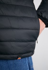 Zwarte pufferjack met horizontale stiksel, elastische manchetten en een ritszak. Voorzien van een klein merklabel nabij de zoom.