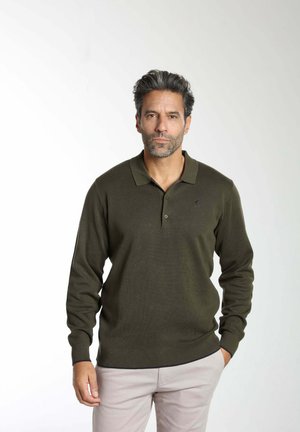 Homme aux cheveux poivre et sel portant un polo vert foncé à manches longues et un pantalon beige clair, debout sur un fond blanc uni.