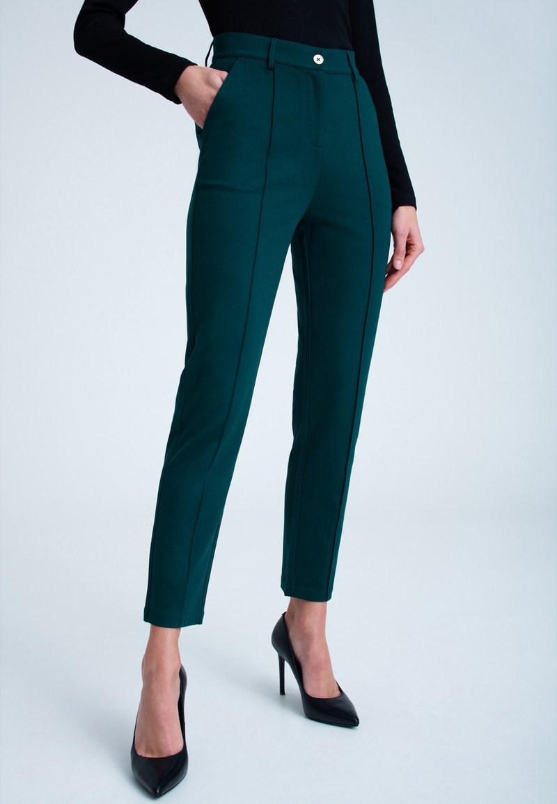 Zielone dopasowane spodnie z pionowymi szwami, z dopasowaną talią i zapięciem na guzik z przodu. Połączone z czarnymi szpilkami.