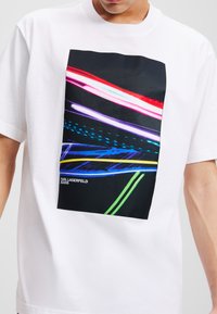 Balta medvilninė marškinėliai su stačiakampiu grafiniu spausdinimu, kuriame yra ryškios, spalvotos šviesos juostos juodame fone. Įtrauktas logotipo detalė.