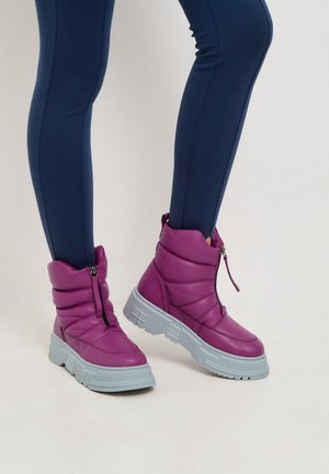 Nogi w dopasowanych granatowych spodniach noszące jaskrawo fioletowe ocieplane buty z przednimi suwakami i grubymi teksturowanymi jasnoszarymi podeszwami na jednolitym tle.