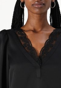 Svart bluse med dyp V-hals som har intrikate detaljer i svart blonder. Glatt stoff, puffede ermer, og en subtil stolpe ved halslinjen.