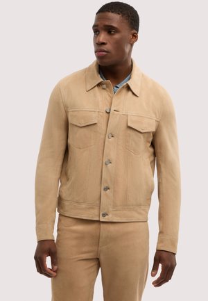 Mand iført beige ruskindjakke med knaplukning og matchende beige bukser, kigger let mod venstre mod en ensfarvet baggrund.