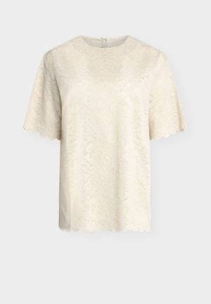 Camicia corta in pizzo color crema con motivo floreale, bordi smerlati e scollo rotondo, esposta su uno sfondo semplice.