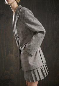 Grijze geruite blazer met brede revers en twee zwarte knopen, gecombineerd met een bijpassende geplooide rok met gelaagde franjes.