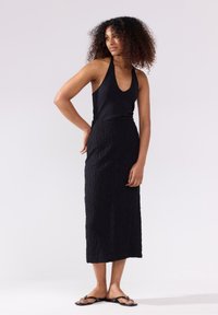 Top senza maniche nero con scollatura a V profonda e gonna nera testurizzata fino al pavimento. La modella indossa sandali neri flat. Sfondo neutro.