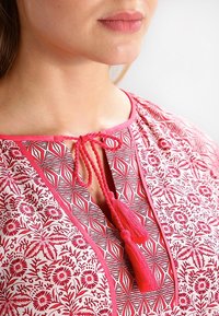 Tuniková halenka s květinovým vzorem v červené a bílé barvě, s kulatým výstřihem se šnůrkami a třásněmi, krátkými rukávy a složitou texturou látky.