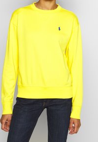 Žlutá mikina z měkké látky s kulatým výstřihem a žebrovanými manžetami. Na levé straně hrudi je malý modrý nápis. Kombinováno s tmavými džínami.