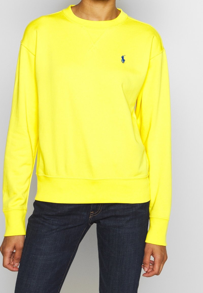 Žlutá mikina z měkké látky s kulatým výstřihem a žebrovanými manžetami. Na levé straně hrudi je malý modrý nápis. Kombinováno s tmavými džínami.