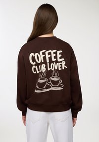 Osoba ubrana w brązową bluzę z napisem "KONESER KAWY" oraz grafiką dwóch parujących filiżanek kawy na plecach, w połączeniu z białymi spodniami.