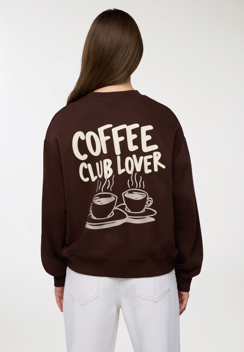 Osoba ubrana w brązową bluzę z napisem "KONESER KAWY" oraz grafiką dwóch parujących filiżanek kawy na plecach, w połączeniu z białymi spodniami.