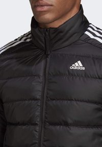 Gros plan sur une personne portant une doudoune noire Adidas avec des bandes blanches sur les épaules et une fermeture éclair sur le devant.