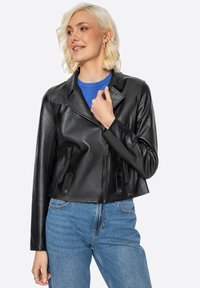 Schwarze Leder-Motorradjacke im cropped Stil mit einem geschlitzten Kragen, Reißverschluss vorne und zwei seitlichen Taschen. Getragen über einem blauen Pullover.