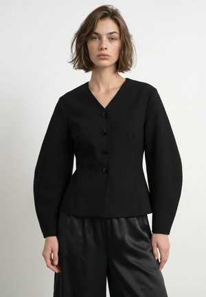 Femme aux cheveux courts et bruns, vêtue d'une veste noire ajustée boutonnée à manches longues et d'un pantalon en satin noir, sur un fond clair uni.