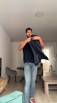 Homme en débardeur noir et jean en train d’enfiler une veste sombre dans un salon avec un mobilier moderne et un sol en bois.