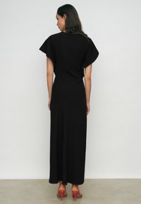 Fekete maxi ruha fodros ujjakkal, ívelt varrási részletekkel és kontrasztos varrással. Narancssárga magassarkú szandállal viselve.