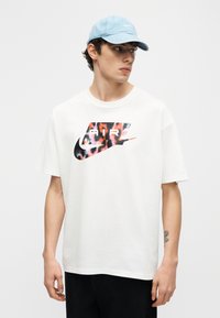 Jauns vīrietis valkā baltu Nike Air grafisko t-kreklu, gaiši zilu cepuri un melnas bikses, skatoties uz kreiso pusi pret vienkrāsainu fonu.