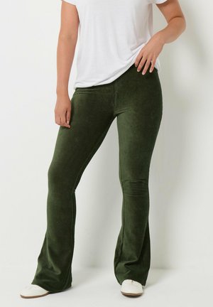 Persoon draagt groene corduroy flare broek en witte schoenen, staat ontspannen met één hand in de zak en de andere op de heup.