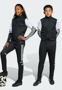 Czarne dresy sportowe z białymi akcentami i trzema paskami na rękawach oraz nogawkach. Model w czarnych butach piłkarskich, trzymający piłkę nożną.